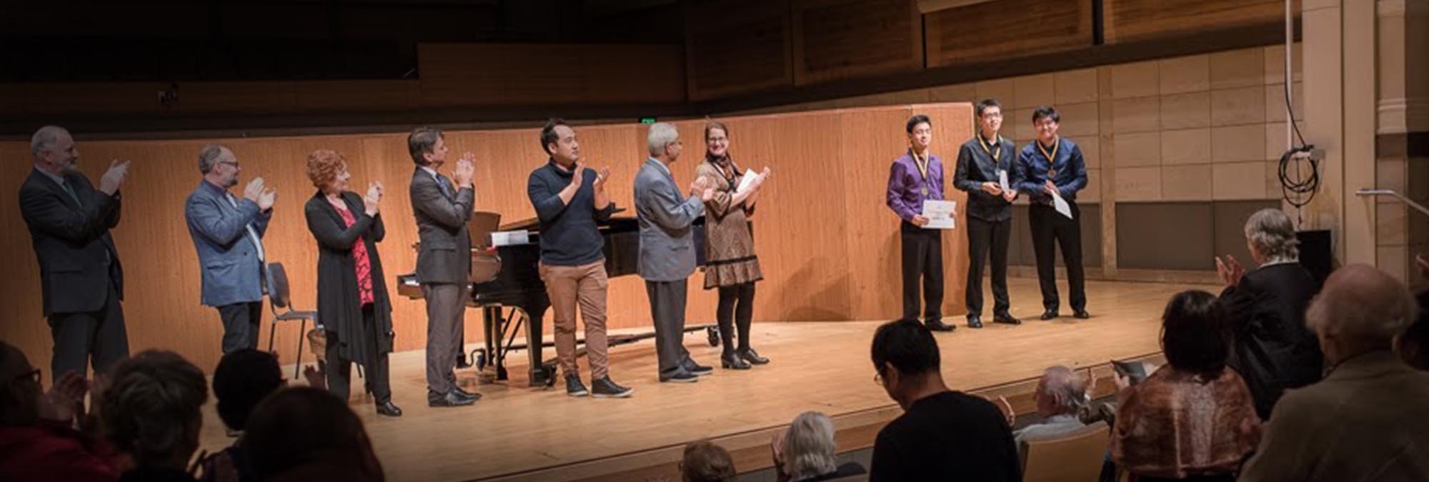 Seven older individuals on a stage applauding for what appears to be three younger award winners