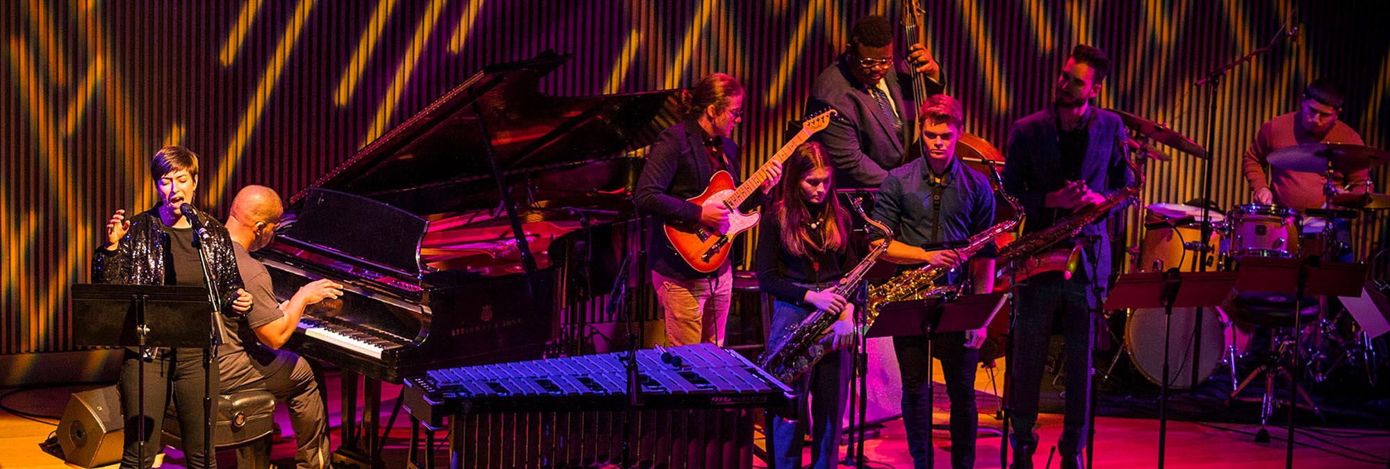 Seven individuals on a stage in a jazz combo setup, guitars, voice, piano, with vibrant lighting