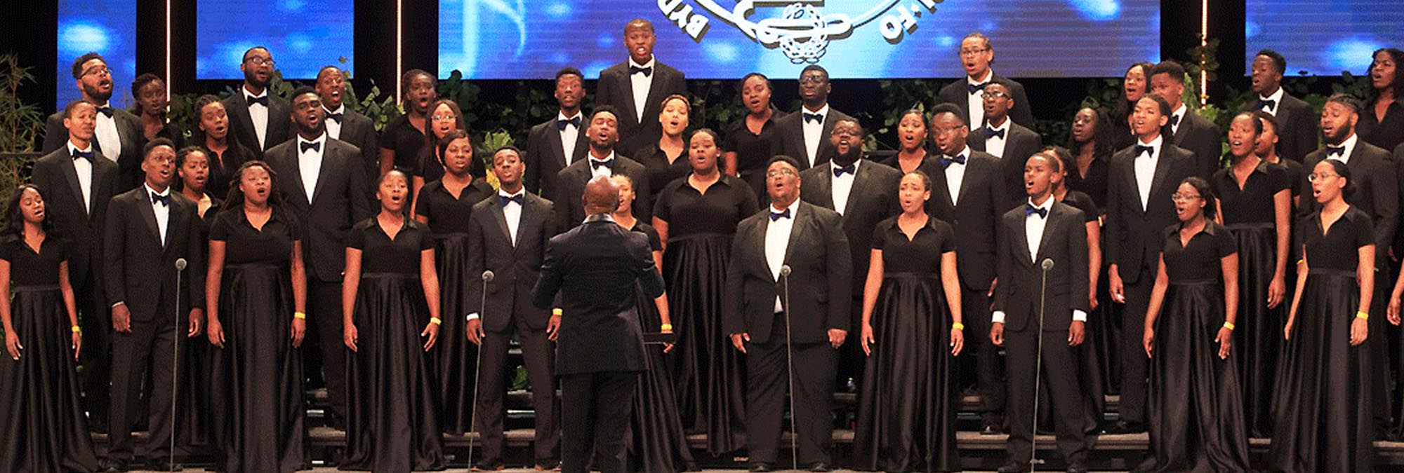 Many individuals, dressed in plain black clothing, being directed in song on a stage as a part of a choir