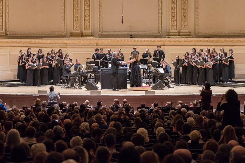 An ensemble of singers and instrumentalists standing onstage in front of a full concert hall