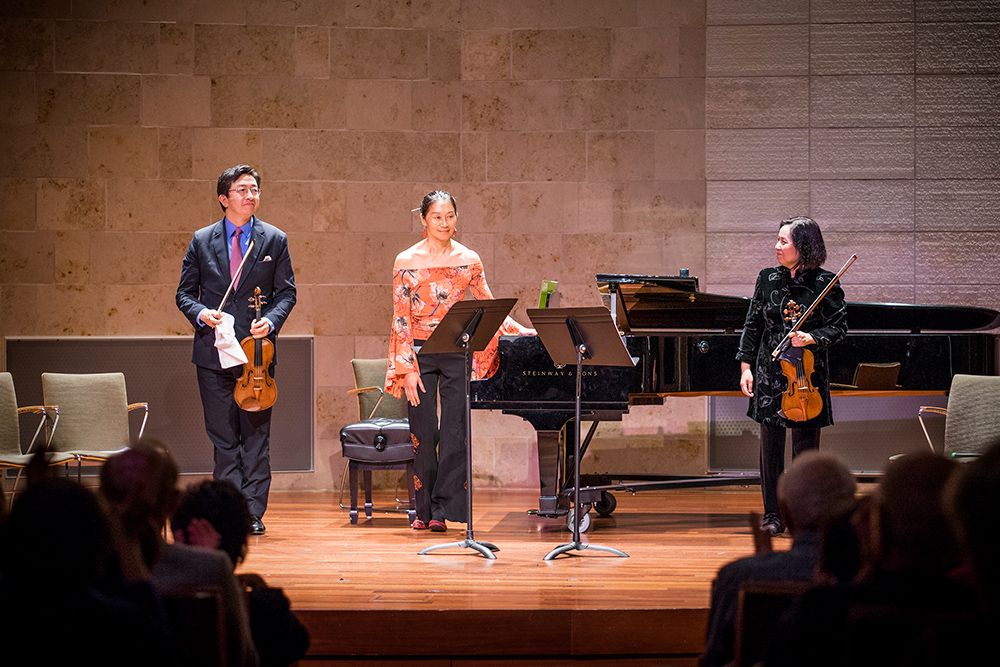 Two violinists and a pianist stand on stage in the Recital Hall before a performance