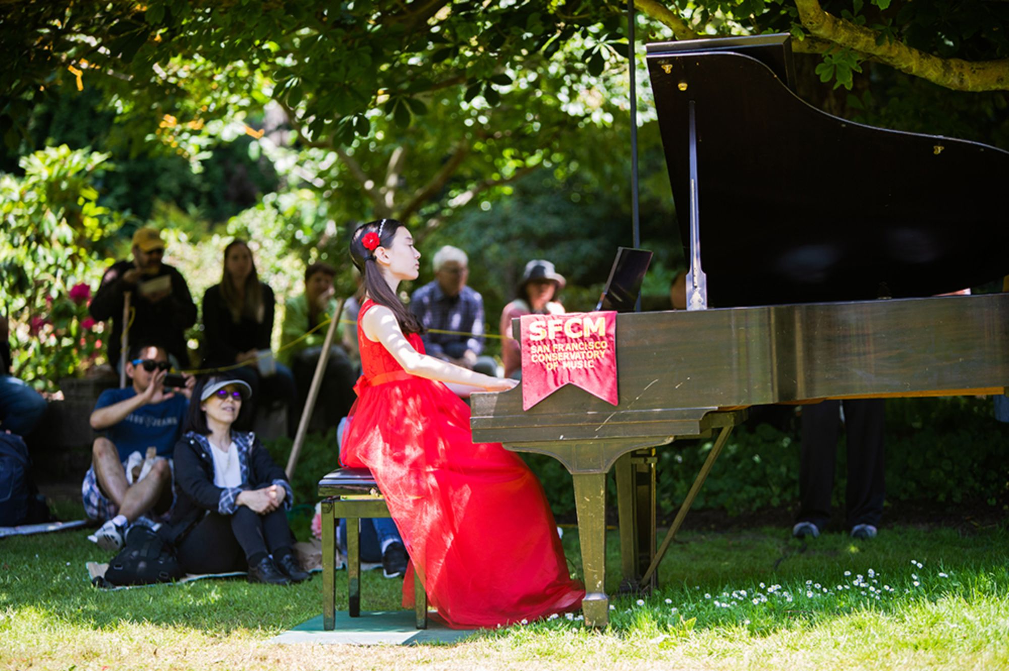 A summer pre-collage student plays the piano at an outdoor concert.  She is wearing a red dress and is surrounded by a small crowd of people smiling at her performance