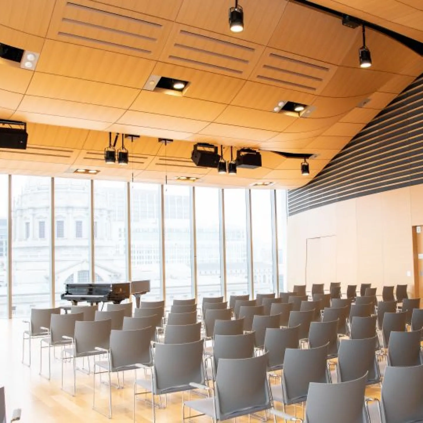 a piano on stage in the osher recital hall. there are lots of windows and city hall is in the background