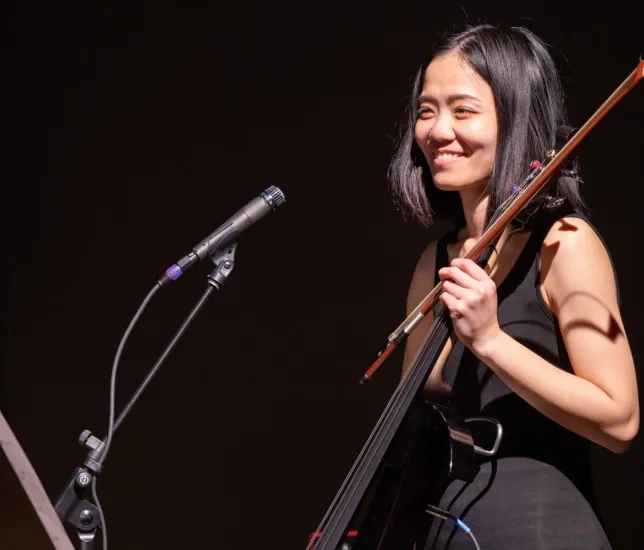 a student smiles holding their electric cello in front of a microphone