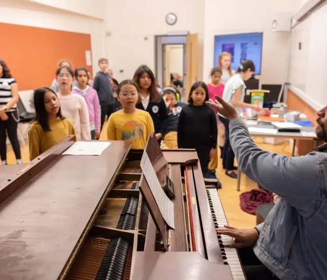 Young students gather around a teacher seated at a piano.