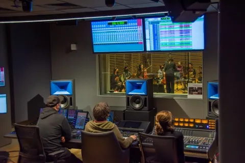 people sit in the recording booth of studio h while looking into studio g and the performers inside