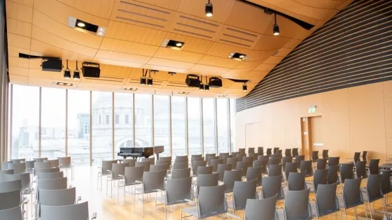 a piano on stage in the osher recital hall. there are lots of windows and city hall is in the background