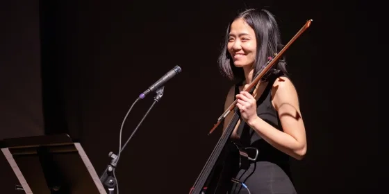 a student smiles holding their electric cello in front of a microphone