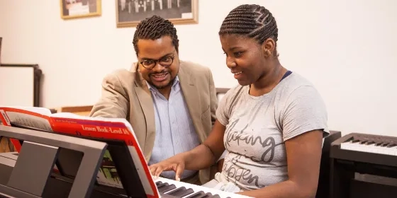 A male teacher with a female student at a keyboard with the teacher pointing to some sheet music