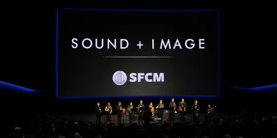A large, dark room with a huge screen with the text, "Sound + Image" and a brass brand before it