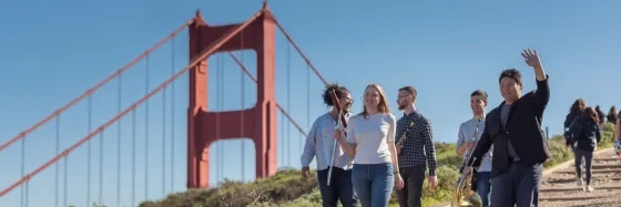 students hold their instruments and walk along a trail where the tops of the golden gate bridge is visible behind them