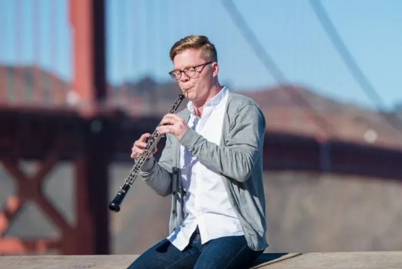 an oboe player sits on a ledge while playing the oboe in front of the golden gate bridge