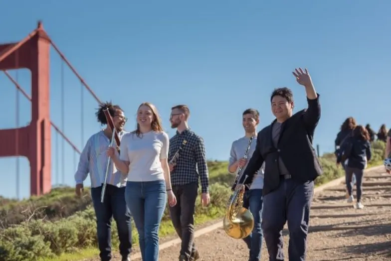 students smile and wave on a trial in front of the golden gate bridge holding their instruments
