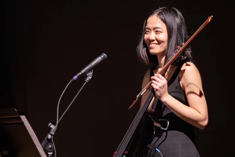 a student smiles holding their electric cello in front of a microphone