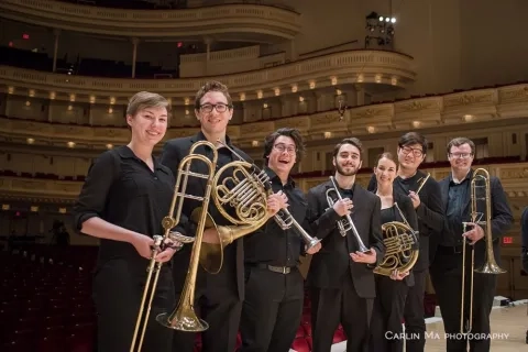 Seven musicians smiling and holding their instruments on the stage of a concert hall