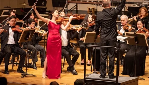 a solo violinist in a red dress plays in front of an orchestra