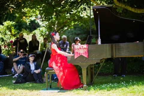 A summer pre-collage student plays the piano at an outdoor concert.  She is wearing a red dress and is surrounded by a small crowd of people smiling at her performance