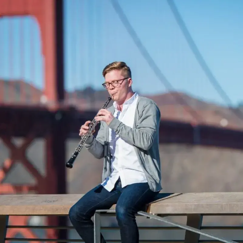 an oboe player sits on a ledge while playing the oboe in front of the golden gate bridge