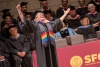 A woman wearing a graduation cap and gown with a rainbow stole smiles with her arms in the air