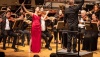 a solo violinist in a red dress plays in front of an orchestra
