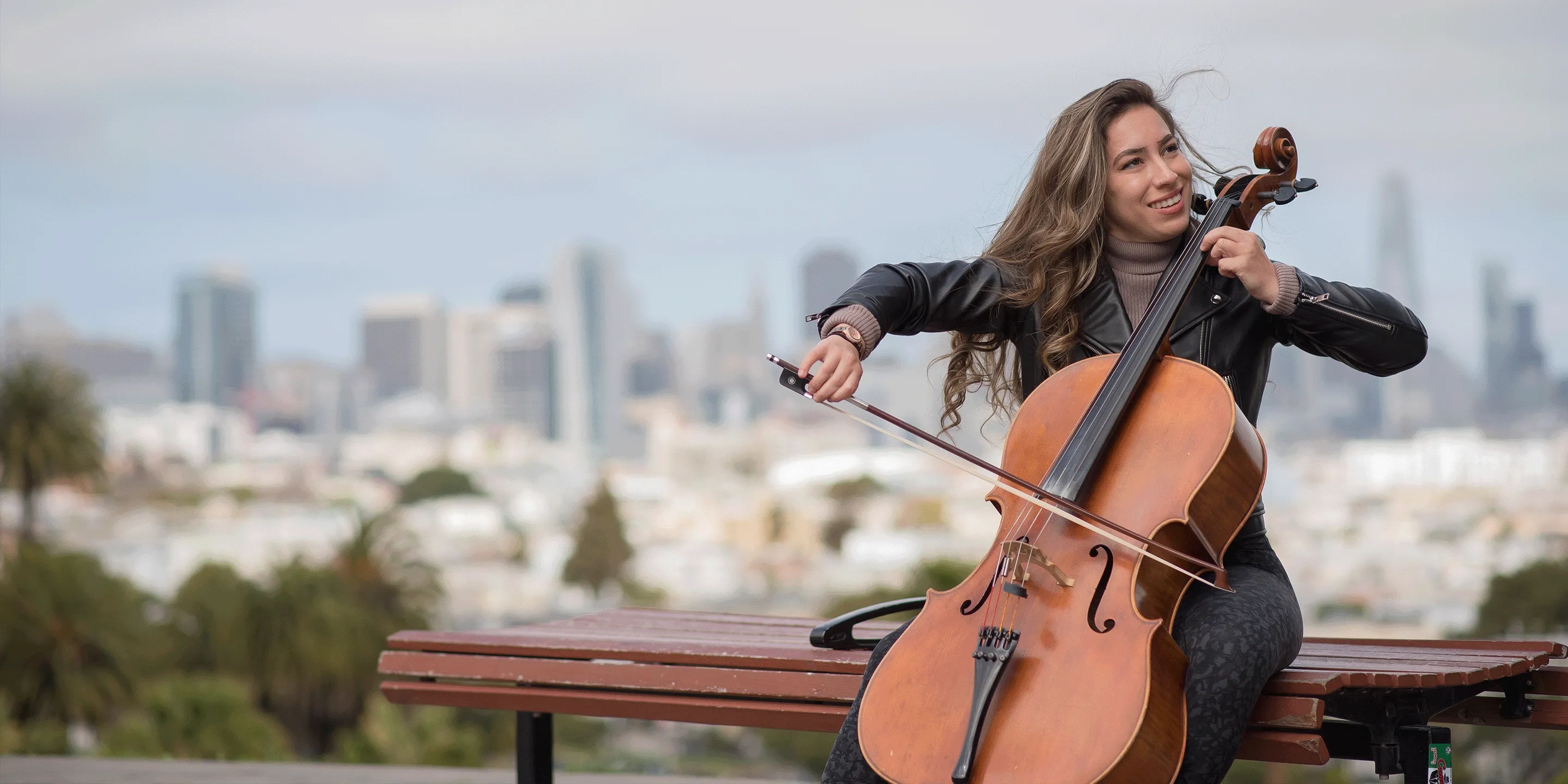 A student plays cello on a park bench with the San Francisco skyline behind her.